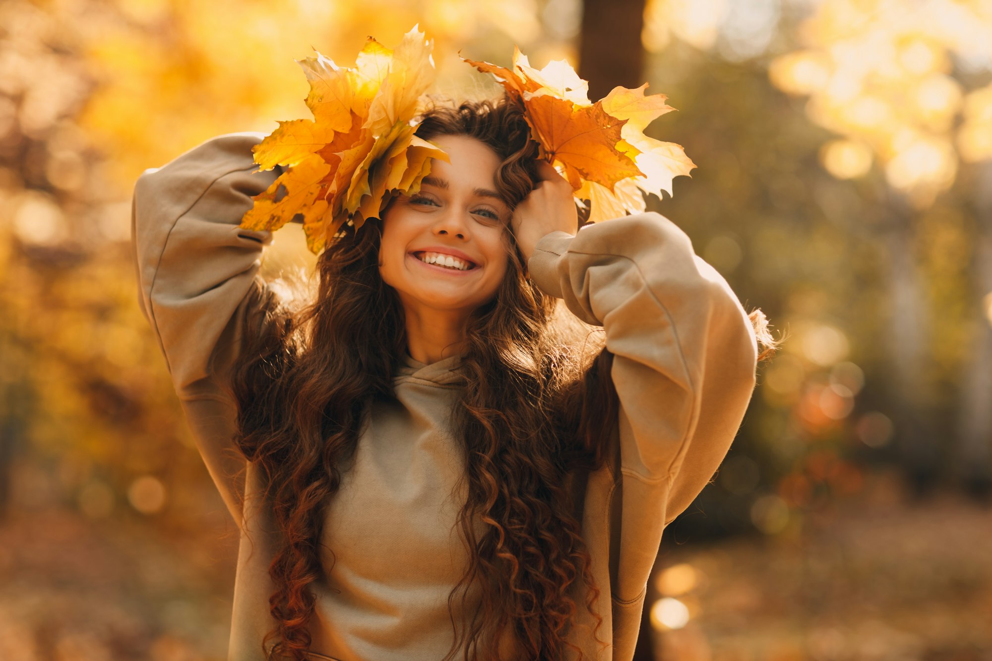 Smiling young woman enjoys the autumn weather in the fall park with the yellow leaves at sunset